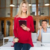 Frau steht mit Ihrem Kollegen im Büro. Sie hält ein Tablet in der Hand, der Mann arbeitet am Laptop.