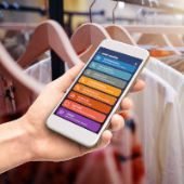 Eine Hand hält ein Smartphone fest mit geöffneter App und im Hintergrund sind Blusen auf Kleiderbügeln an einer Stange zu sehen