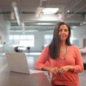 Frau steht selbstbewusst in Ihrem Unternehmen. Neben ihr steht auf einem Tresen ein Laptop.