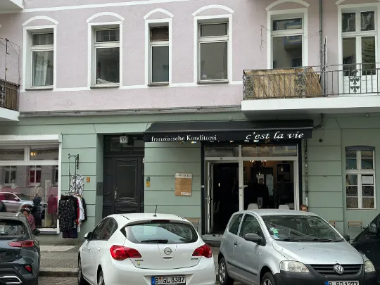 Ein kleines, französisches Café in Berlin Friedrichshain. Davor stehen geparkte Autos. Über dem Café ist eine hübsche Wohnung mit Balkon zu sehen.