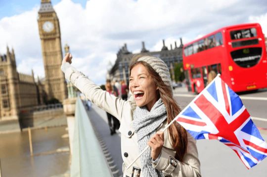 Abitur-Absolventin steht in London vor dem Big Ben mit einer UK-Flagge in der Hand und freut sich über ihr gutes Englisch